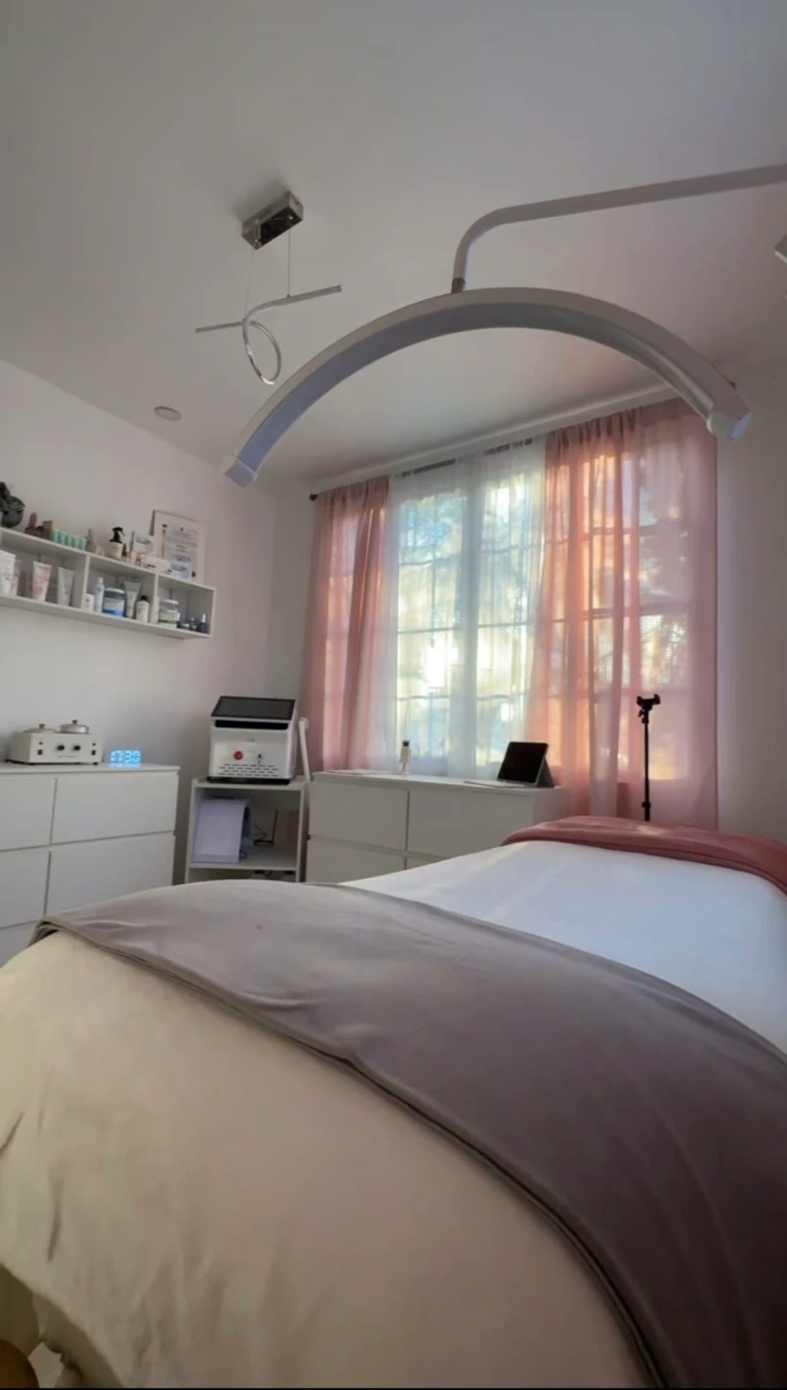 La cabina del estudio: camilla lista bajo la lámpara de arco, cortinas rosadas y luz de ventana