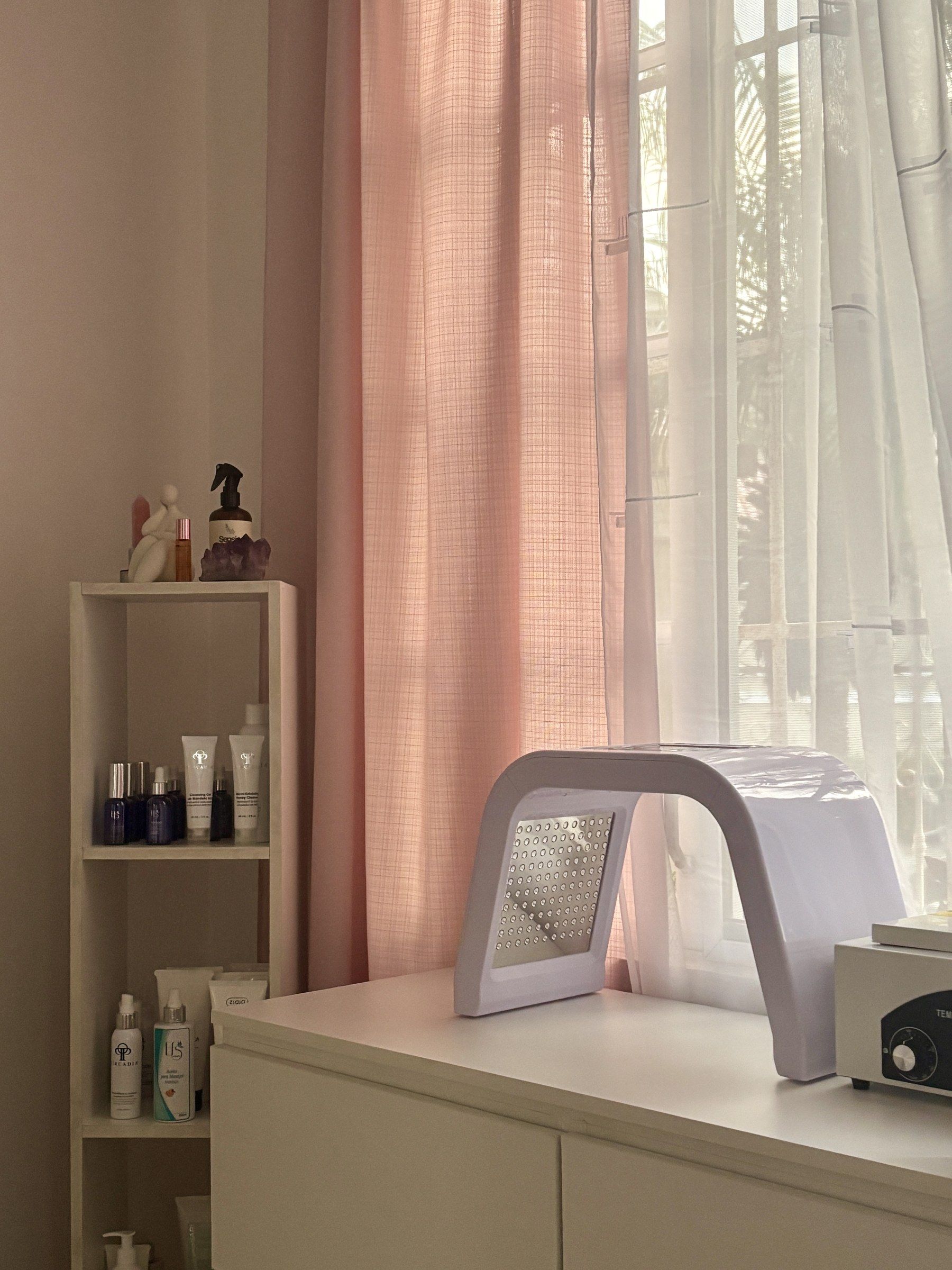 La cabina vacía y ordenada: estante con productos profesionales y el equipo de luz LED sobre la mesa, junto a las cortinas rosadas