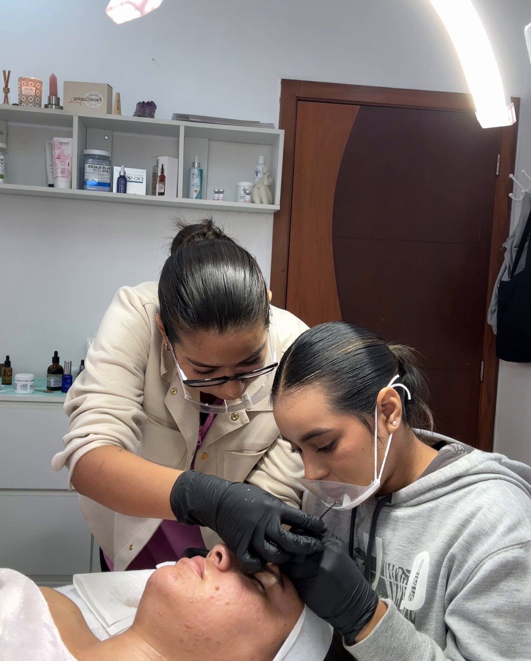 Ambar, con chaqueta beige y lentes, guía de cerca a una alumna que trabaja con guantes sobre el rostro de una modelo